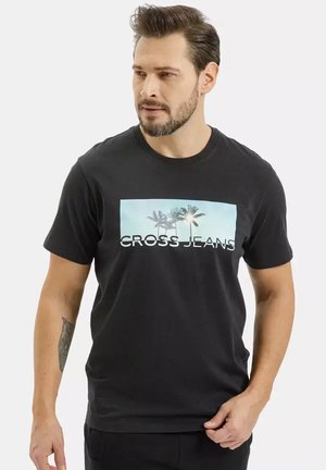 Mężczyzna z krótkimi ciemnymi włosami i brodą, ubrany w czarną koszulkę z motywem palm i napisem "CROSS JEANS", patrzy lekko w lewo.