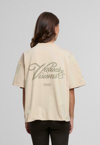 Beige oversized t-skjorte med korte ermer, med et grønt grafisk trykk på ryggen som sier "Verdier & Visjoner" og "Pegador."