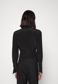 Černá saténová blůza s přiléhavým střihem, dlouhými rukávy a knoflíkovými manžetami, a detailním skladem na zadní straně. Hladká textura a lesklý povrch.