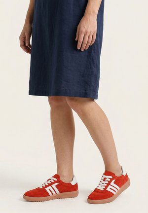 Person iført en knælang navy kjole og røde sneakers med hvide striber og gummisåler, stående på en ensfarvet lys baggrund.