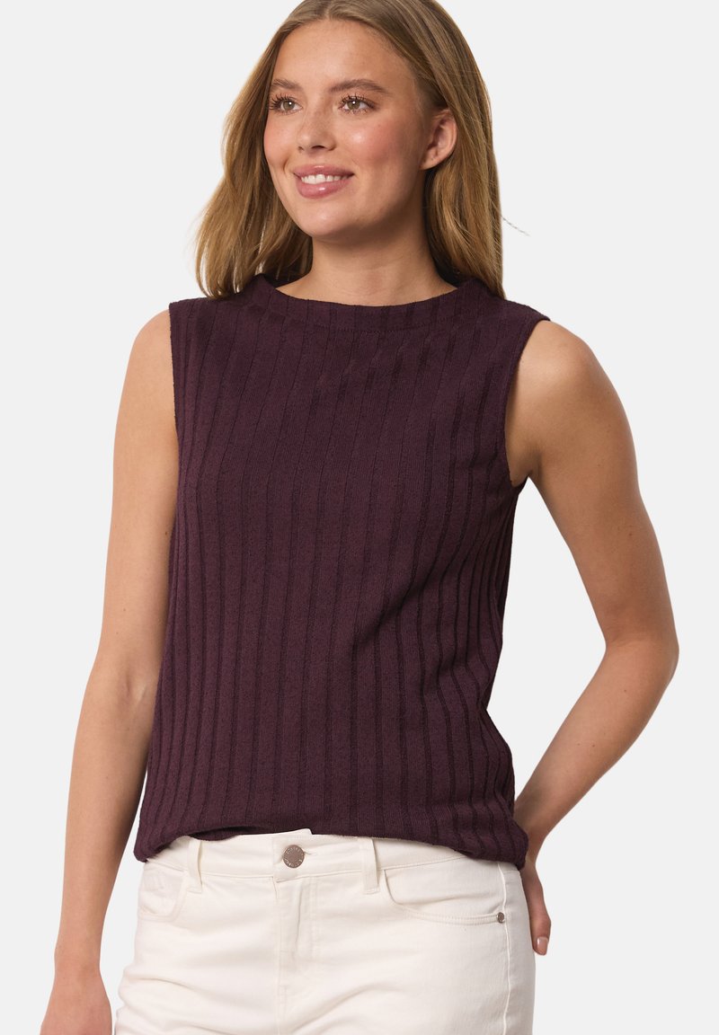 Jeune femme souriante, portant un top violet sans manches en côtes et un pantalon blanc, debout avec une main derrière le dos.