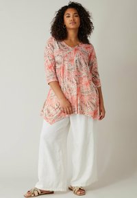 Tunika a fantasia corallo e beige con maniche 3/4, scollo a V e una forma fluida, abbinata a pantaloni larghi in lino bianchi e sandali beige.