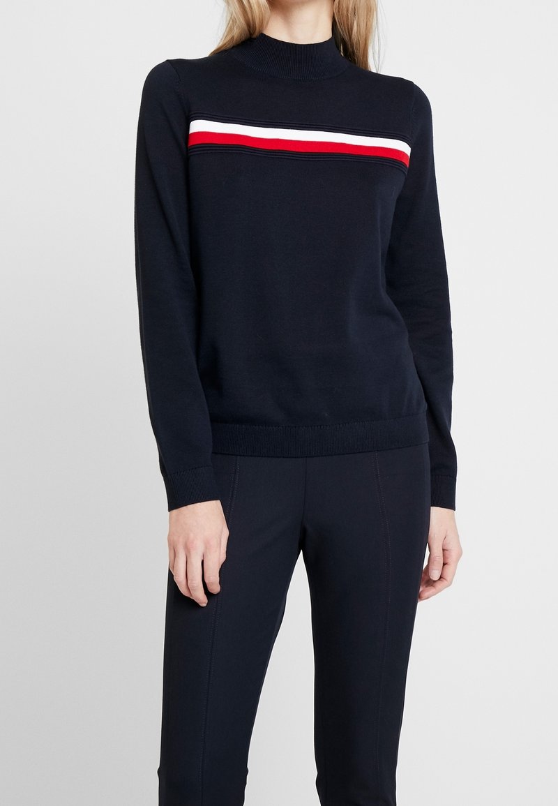 Femme portant un pull à manches longues bleu marine avec des rayures horizontales rouges, blanches et noires sur la poitrine, associé à un pantalon bleu marine.