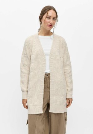 Jeune femme portant un long cardigan en tricot crème, un haut blanc et un pantalon marron ample, debout devant un fond blanc uni.