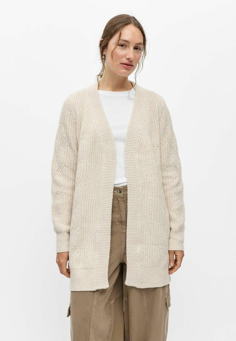 Jeune femme portant un long cardigan en tricot crème, un haut blanc et un pantalon marron ample, debout devant un fond blanc uni.