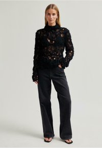 Černý texturovaný svetr s vysokým límcem, s vrstveným designem a otevřeným pletením, spárovaný s černými džínami s vysokým pasem.