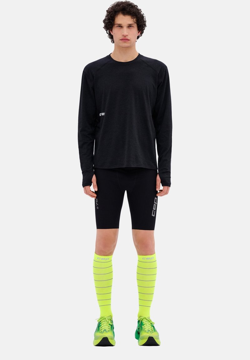 Zwarte long-sleeve shirt, zwarte strakke shorts, geel gestreepte compressiesokken tot aan de knie en groene hardloopschoenen. Logo's zichtbaar op de kleding.