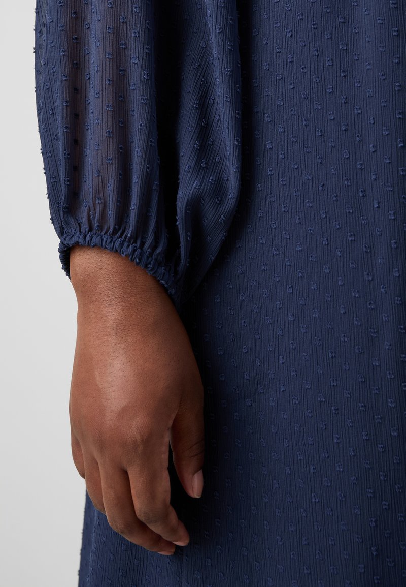 Robe bleu marine en tissu texturé avec de petits points en relief. Manches longues froncées avec des poignets élastiques qui accentuent le design.