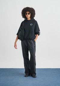 Μαύρο φούτερ με κουκούλα και ελαστική ραφή, μαύρο παντελόνι φόρμας με λευκή ρίγα και μικρή μαύρη τσάντα ώμου. Το μοντέλο φορά γυαλιά ηλίου.