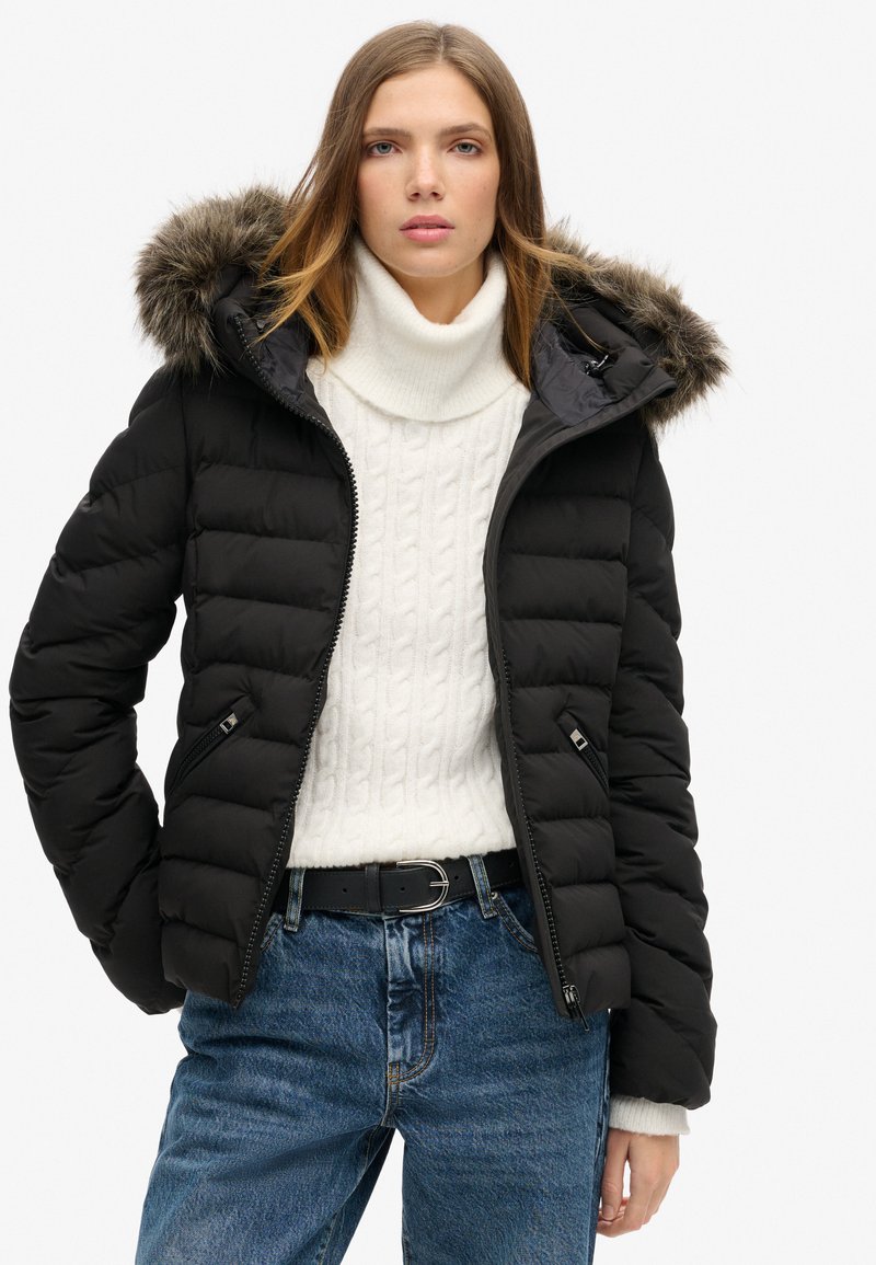 Zwarte pufferjack met een capuchon met bontafwerking, ritssluiting aan de voorkant en zijzakken, gedragen over een witte kabelgebreide trui en blauwe jeans.