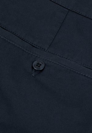 Tessuto blu navy con una trama texturizzata, caratterizzato da un bottone nero e cuciture visibili. Il design include una tasca e dettagli di cucitura diagonale.