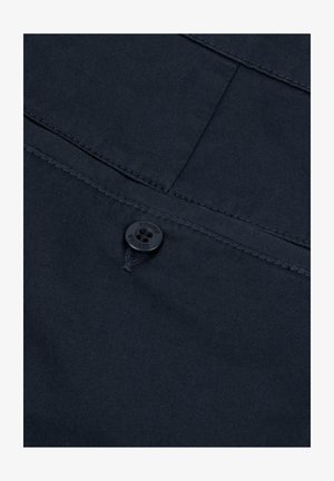 Tessuto blu navy con una trama texturizzata, caratterizzato da un bottone nero e cuciture visibili. Il design include una tasca e dettagli di cucitura diagonale.