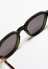 Groene transparante zonnebril met grijze lenzen. Hoekig design en dikke monturen met pootjes in schildpadpatroon. Glad oppervlak.