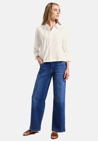 Vit skjorta med långa ärmar, i kombination med vida blå jeans och bruna sandaler. Tyget ser mjukt ut med en avslappnad passform.