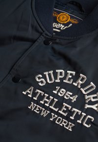 Veste bleu marine avec boutons-pression noirs et texte brodé blanc "Superdry 1964 Athletic New York" sur le devant.
