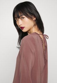 Blusa rosa poeirenta com um delicado detalhe em renda nas costas, corte solto, mangas longas e laços de fita no decote, confeccionada em tecido leve.