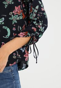 Mörkblå blus med lång ärm och blommiga samt kattmönster, med dragkedjedetalj vid ärmsluten, tillverkad av lättviktigt material.