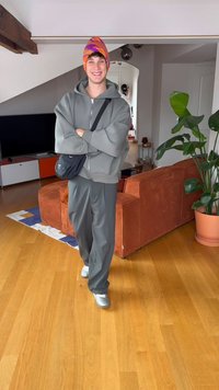 Φούτερ με φερμουάρ γκρι πάνω από γκρι φαρδιά παντελόνια, πορτοκαλί και μωβ μαντήλι με σχέδιο, λευκά αθλητικά παπούτσια, ορθός σε μοντέρνο εσωτερικό χώρο.