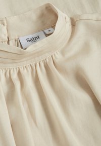 Beige blúz, ami összehúzott nyakkivágással, puha textúrával rendelkezik, és a "Saint Tropez" márkajelzés látható a belsejében. A méretezés szerint közepes.