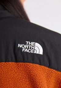Giacca in fleece con un corpo arancione testurizzato e parte superiore in nylon nero. Il logo The North Face è ricamato in bianco sul petto.