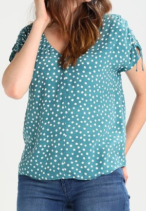 Teal short-sleeve top featuring white polka dots, a V-neckline, and tie details on sleeves. Made from a lightweight fabric with a relaxed fit.