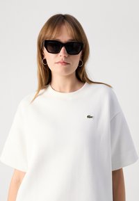 White cotton sweatshirt with short sleeves and a round neckline, featuring a small green embroidered logo on the left chest. Black sunglasses.