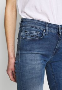 Vaqueros de denim en un degradado azul oscuro, con una silueta ajustada, costuras en contraste y un pequeño bolsillo trasero con un diseño en ángulo.