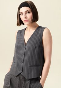 Grå rutiga västen med djup V-hals, fem knappar och sidofickor. Tillverkad av slät tyg, med en skräddarsydd, figurnära design.