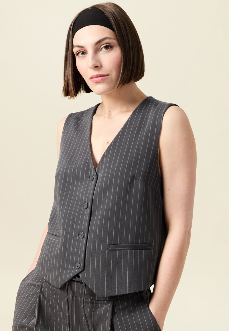 Grå rutiga västen med djup V-hals, fem knappar och sidofickor. Tillverkad av slät tyg, med en skräddarsydd, figurnära design.