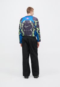 Μπλούζα με μακριά μανίκια και πολύχρωμο γεωμετρικό σχέδιο, με μπλε ώμους και μαύρη βάση, συνδυασμένη με φαρδιά μαύρα παντελόνια.