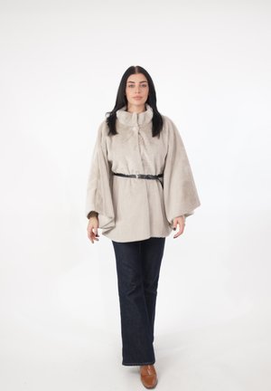 Donna con capelli lunghi e scuri che indossa una giacca di pelliccia beige con cintura nera, jeans scuri e scarpe marroni, in piedi su sfondo bianco.