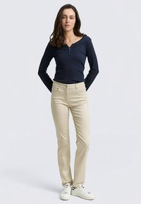 Marineblaues Henley-Oberteil mit langen Ärmeln kombiniert mit beigen, schmal geschnittenen Hosen. Die Hose verfügt über Vordertaschen und einen flachen Bund. Weiße Turnschuhe vervollständigen das Outfit.