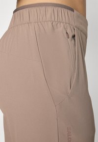 Lichtbruine sportbroek van rekbaar materiaal, met een elastische tailleband en een zijzak met een klein ritsdetail. Logo op het been gedrukt.