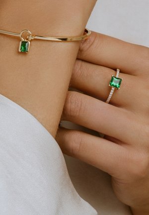 Guldarmbånd med grøn halvædelsten charm og matchende ring. Begge har poleret metal og intrikate stenindstillinger. Glat hudtekstur synlig.