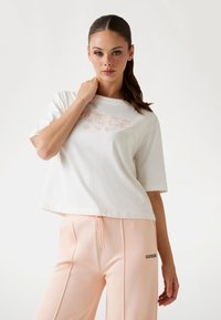 Maglietta bianca corta con logo "GUESS" in rosa chiaro, abbinata a pantaloni tuta pesca con un logo nero sul lato. Texture in cotone morbido.