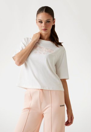 White cropped t-shirt with "GUESS" logo in light pink, paired with peach sweatpants featuring a black logo on the side. Smooth cotton texture.