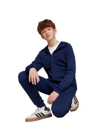 Marineblauwe trainingsoutfit gemaakt van soepel materiaal, met een hoodie met rits en smalle broek. Witte t-shirt eronder, gecombineerd met zwart-witte sneakers.
