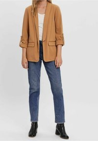 Béžové sako s ohrnutými rukávy, dvěma předními kapsami a volným střihem, doplněné tmavě modrými džíny s přímými nohavicemi a černými kotníkovými botami.