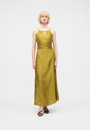 Γυναίκα με σγουρά ξανθά μαλλιά που φοράει αμάνικο μάξι φόρεμα ελιάς και μαύρες ψηλοτάκουνες σανδάλες με λουράκια, στέκεται μπροστά σε ένα απλό λευκό φόντο.