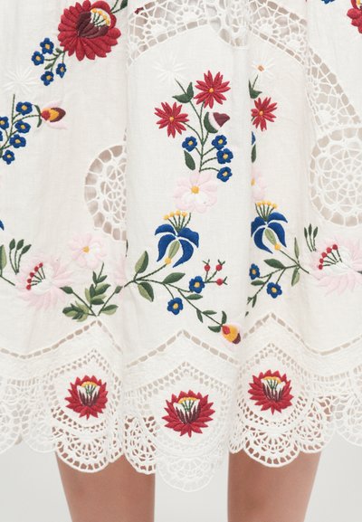 Besticktes weißes Kleid mit bunten Blumenmustern in Rot, Blau und Pink. Eine Spitzenkante am Saum verleiht ihm komplexe Textur und Detail.