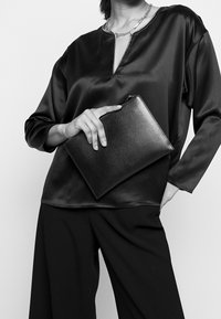 Haut en satin noir avec un col en V et des manches longues, associé à un pantalon noir à jambes larges, tenant une pochette noire texturée avec une fermeture éclair.