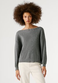 Pelēka džemperis ar adījuma struktūru, plašu kakla izgriezumu un smalkiem metāliskajiem caurumiem. Garas, nedaudz pūkainas piedurknes, apvienotas ar gaišām biksēm.