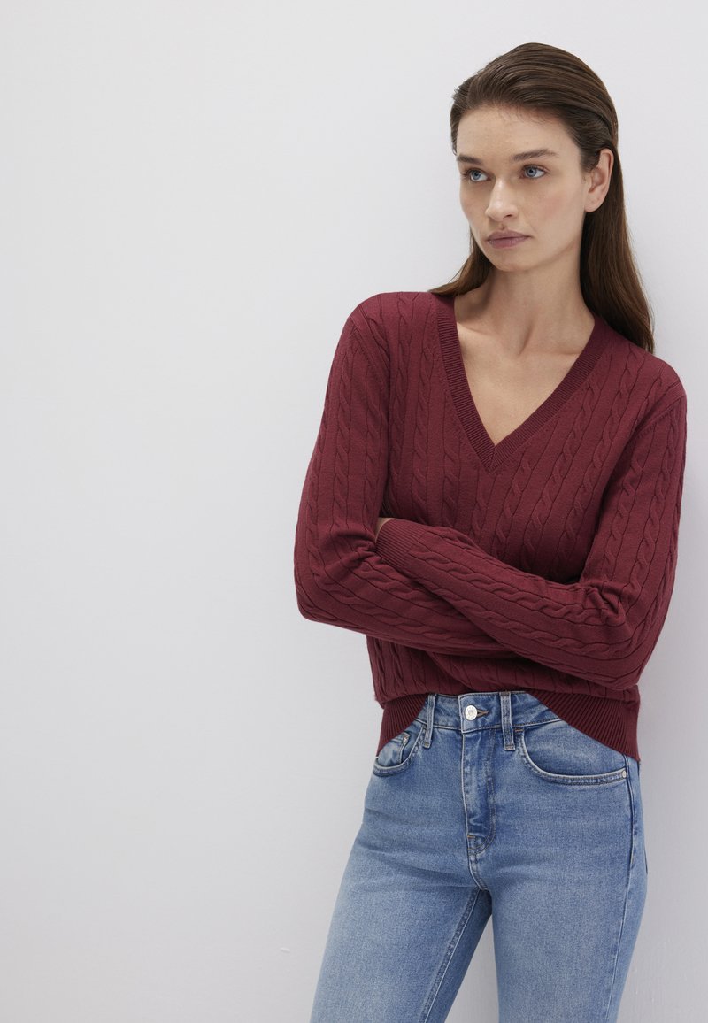 Bordeauxfarbener Strickpullover mit tiefem V-Ausschnitt und gerippten Bündchen, kombiniert mit hellblauen High-Waist-Jeans. Glatte Textur und taillierte Passform.