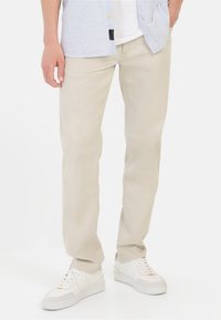 Beige Baumwollhosen mit geradem Schnitt, ausgestattet mit Gürtelschlaufen und Taschendetails, kombiniert mit weißen Sneakers und einem hellblauen Hemd mit Knopfleiste.