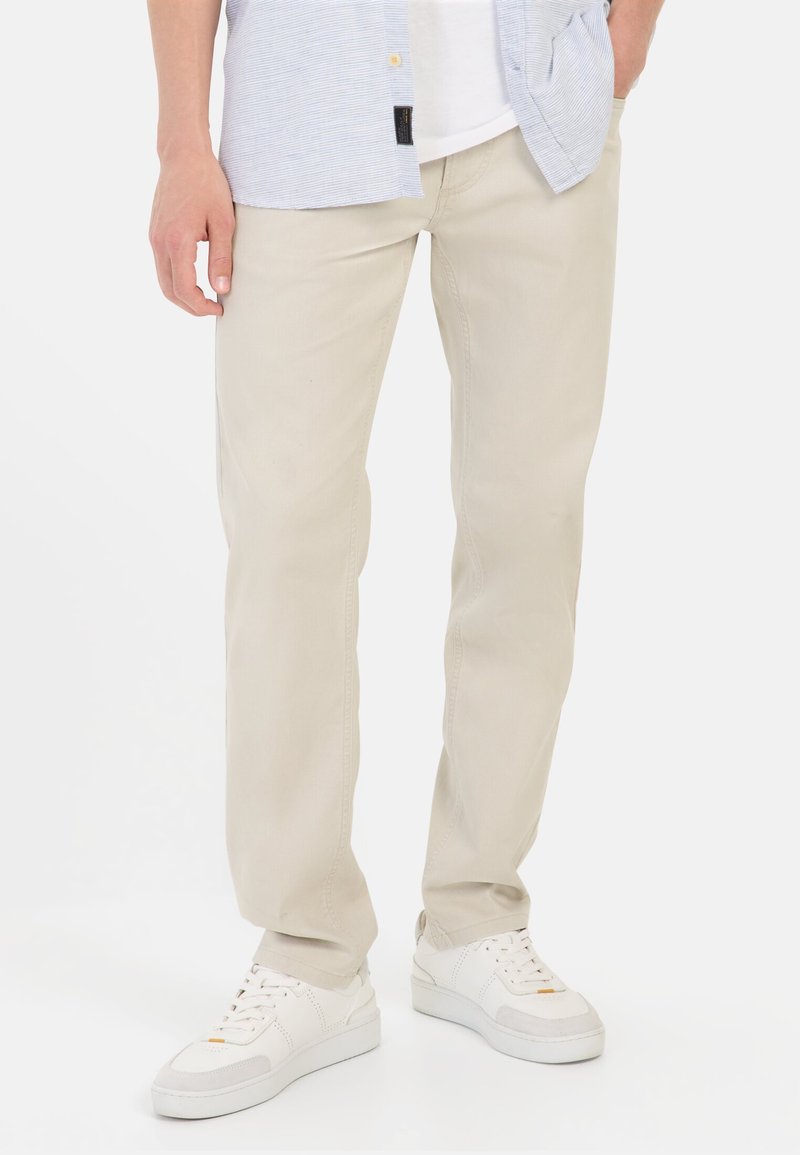 Beige Baumwollhosen mit geradem Schnitt, ausgestattet mit Gürtelschlaufen und Taschendetails, kombiniert mit weißen Sneakers und einem hellblauen Hemd mit Knopfleiste.