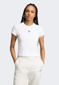 Biely bavlnený skrátený tričko s čiernym logom Adidas na prsiach. Model ho nosí s béžovými pásikavými nohavicami, ruky v vreckách.