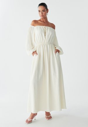 Model wearing an off-the-shoulder, white maxi dress with a cinched waist and long sleeves, standing with hands in pockets.