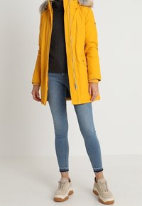 Gul puffjacka med pälskantad huva, dragkedjestängning och fickor, kombinerad med mörk tröja, blå tajta jeans och beige sneakers.