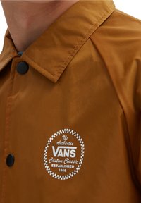 Brown waterproof jacket with a snap button closure and a rounded collar. Features white logo in a circular design on the front.