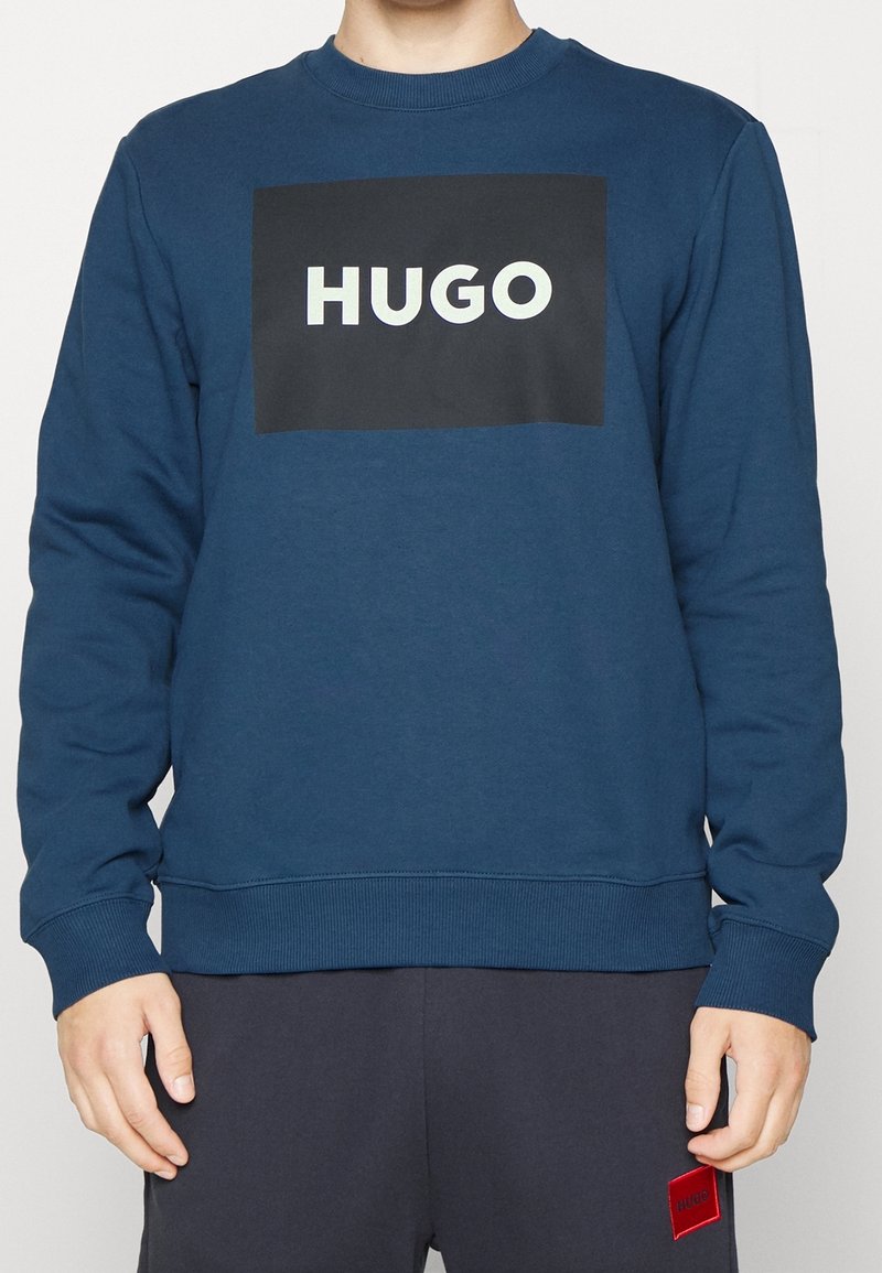 Personne portant un sweat bleu avec "HUGO" en lettres blanches sur un rectangle noir au niveau de la poitrine, associé à un pantalon foncé.